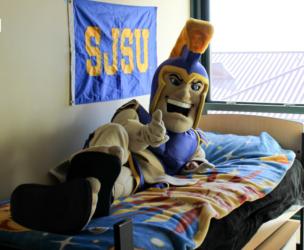 mascot in bed giving thumbs up with 'SJSU' flag in background