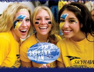 three smiling fans in yellow with face paint holding a 'skyhawk fan' sign