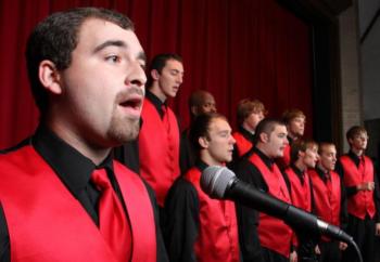 men's choir in black and red attire singing on stage with a microphone