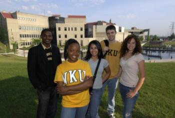 four students smiling with 'nku' shirts in front of campus building