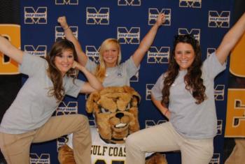 three people posing with a mascot in front of a logo wall