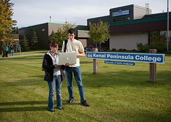 two students with laptop by 'kenai peninsula college' sign